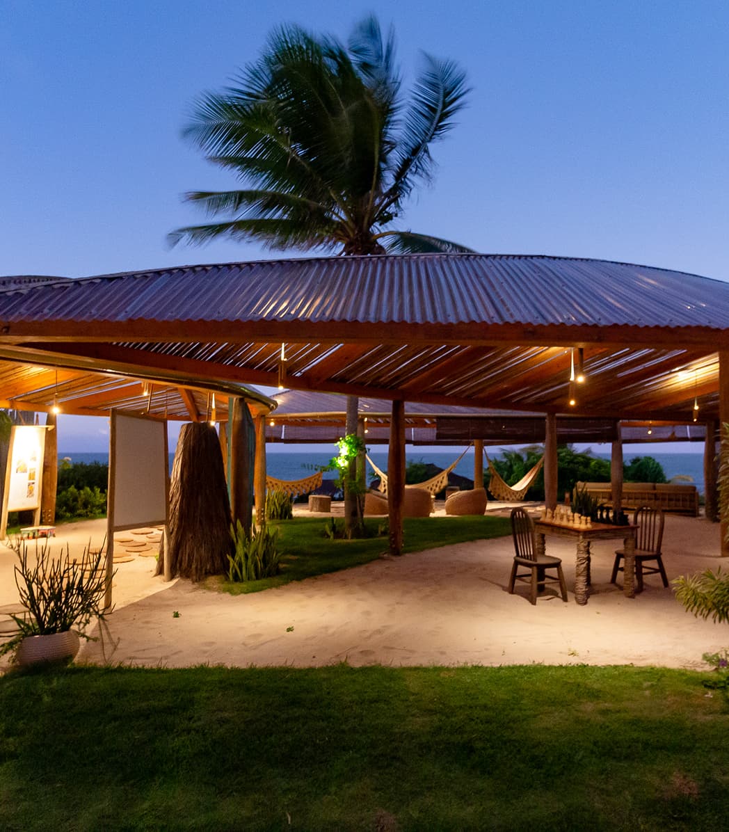 Pavilhão de palha com redes e mesa de madeira iluminados ao entardecer, sob coqueiro, no Kilombo Villas