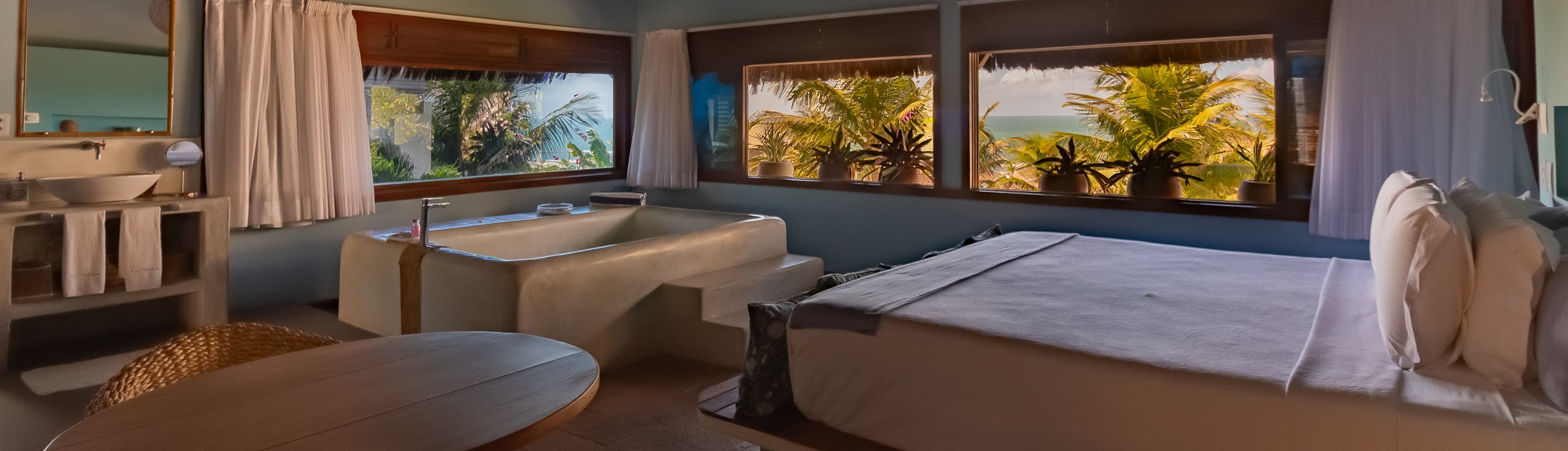 Suíte de spa com maca de massagem e cama de casal diante de janelas com vista para os coqueiros e o mar no Kilombo Villas
