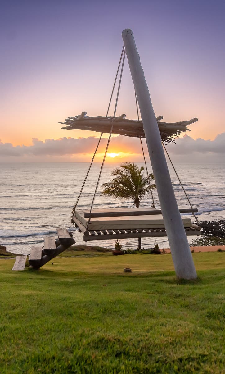 Balanço suspenso de madeira no gramado à beira-mar durante o pôr do sol no Kilombo Villas