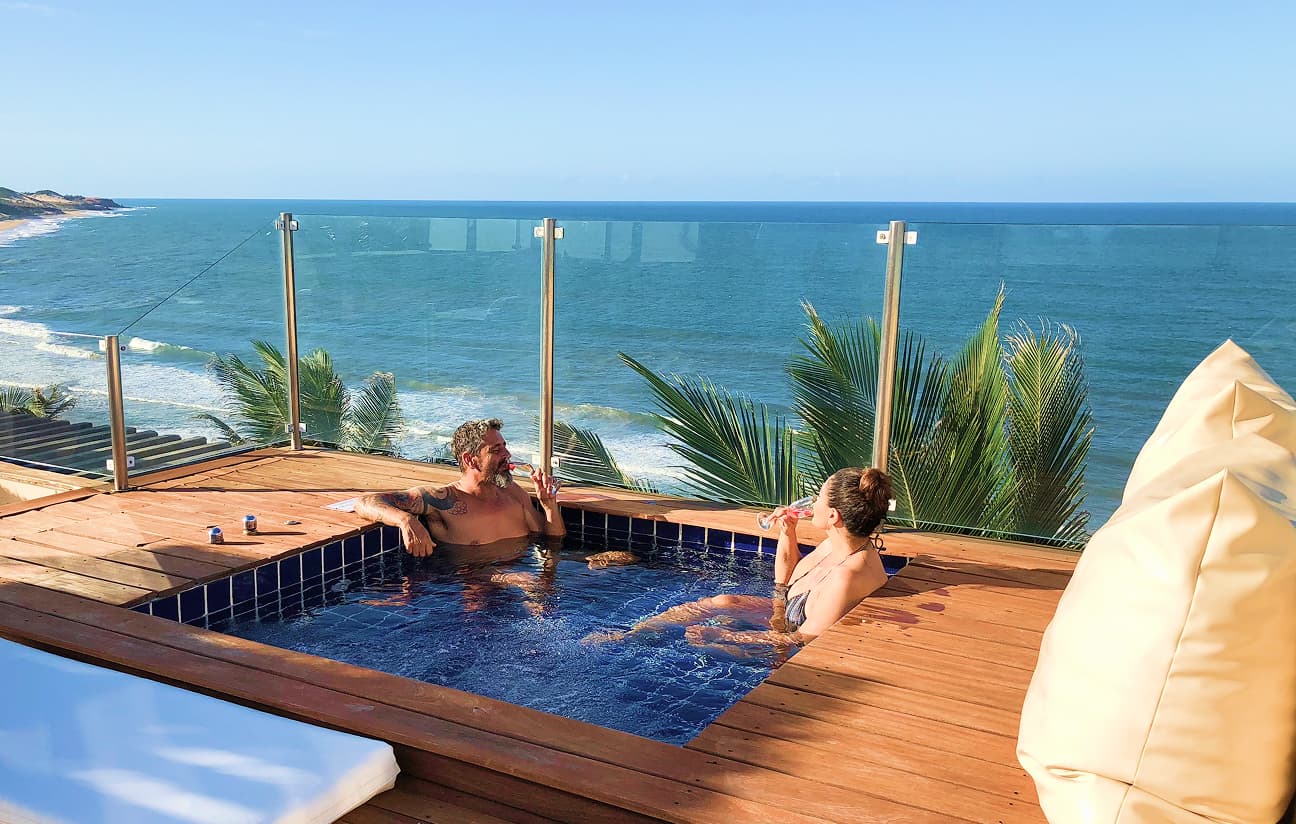 Casal brinda com taças em uma piscina privativa de um deck elevado, com vista panorâmica para o mar e coqueiros.