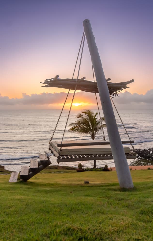 Balanço suspenso de madeira ao pôr do sol com vista para o mar e palmeiras no Kilombo Villas, Sibaúma