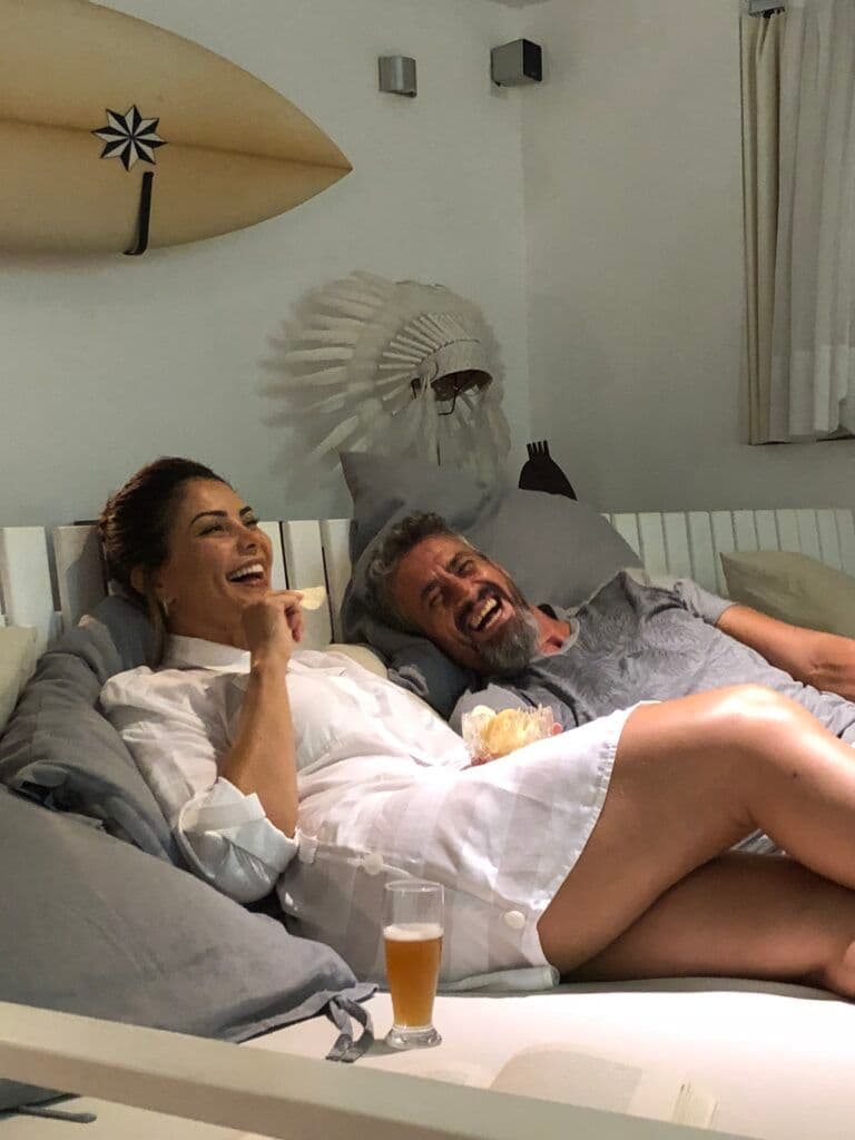 Casal rindo na cama com prancha de surf decorativa, escultura de cocar e cerveja gelada