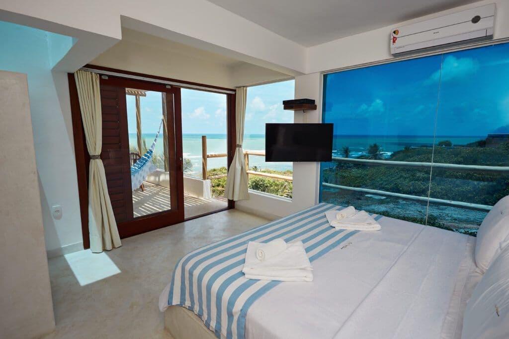 Quarto com cama listrada azul e branca, toalhas dobradas e vista para varanda com rede e oceano