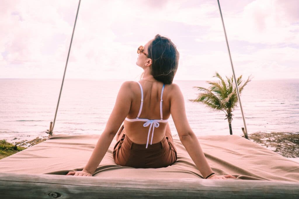 Mulher de biquíni de costas sentada em cama suspensa com óculos de sol e oceano ao fundo